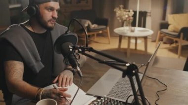 Evde podcast kaydederken loş ışıklandırmalı ve mikrofona konuşan sakallı bir adamın rahat oturma odasında oturduğu el kameralı yavaş çekim.