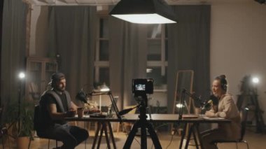 Oturma odasındaki masada oturan genç adam ve kadının stüdyonun ışıkları ve podcast ya da internet şovu ile ışıklandırılmış halleri.