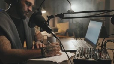 Oturma odasındaki loş ışıklı masada oturan ve mikrofona konuşurken senaryosu hakkında notlar alan sakallı bir adamın görüntüsü.