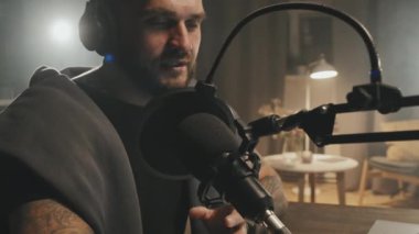 Şapkalı ve kulaklıklı sakallı bir gencin loş ışıklı oturma odasında mikrofona konuşurken podcast veya radyo yayını yaparken görüntüsünü kaldır.