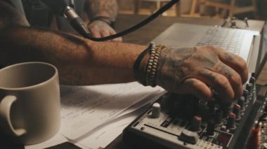 Tanımlanamayan erkek podcast sunucusunun yakın çekim görüntüsü. Dövmeleri var, ses karıştırıcı kullanıyor ve şovunu kaydediyor.