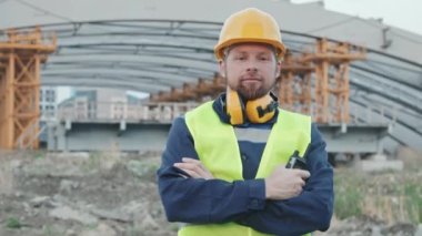 Güvenlik yeleği ve kasketli neşeli erkek inşaat işçisinin portresi kollarını kavuşturup dışarıdaki kameraya bakıyor.