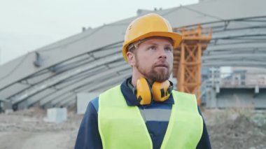 Sakallı erkek inşaat alanının yavaş çekim görüntüleri. Başlık ve güvenlik yeleği giymiş ustabaşı etrafa bakıyor ve iş yerinde dışarıda yürüyor.