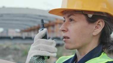 Kadın inşaat işçisi, güvenlik yeleği, kasket ve eldivenlerle dışarıda telsize konuşurken yakın çekim.