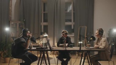 Üç erkek ve kadın podcast sunucusunun loş aydınlatılmış oturma odasında ya da stüdyoda otururken ve mikrofonlara konuşurken görüntülerini izliyoruz.