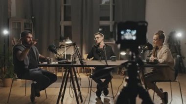 Oturma odasındaki loş ışıklı masada oturan ve podcastlarını kaydederken mikrofonlara konuşan kadın ve erkeklerin görüntülerini izlemek. Video kamera tripodun üzerinde duruyor ve onları çekiyor.