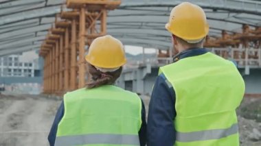 Erkek ve kadın inşaat şantiyesi yöneticilerinin güvenlik yelekleri ve kalın şapkalı dikiz görüntülerinin takibi tamamlanmamış bina veya köprüye doğru yürümeleri ve iskele ile sohbet etmeleri.