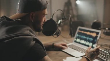 Masada oturmuş, mikrofona konuşan sakallı adamın kadın sunucuyla podcast yaparken çekilen görüntüsü.