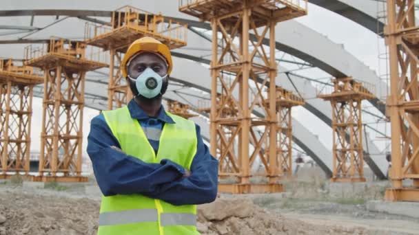 安全ベストのアフリカ系アメリカ人男性建設労働者の追跡肖像画 足場と未完成の建物の前にカメラのポーズハード帽子とほこり顔のマスク — ストック動画