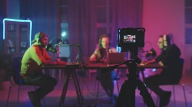 Tripodun üzerinde duran ve oturma odasındaki masada oturan üç kişiyi neon ışıklarla aydınlatan ve podcast kaydeden video kameranın görüntüsü.
