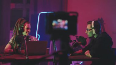Rastalı ve gözlüklü bir adamın neon ışıklarla aydınlatılmış oturma odasında oturmuş podcast kaydeden neşeli genç bir kadının görüntüsü.