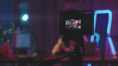 Video kameranın odak noktası tripodun üzerinde dururken yakın çekim yapmak ve oturma odasında oturan üç genç insanı neon ışıklarla aydınlatıp podcast kaydederek çekmek.