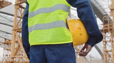 Tanımlanamayan siyah erkek inşaat işçisinin orta kesiminde, güvenlik yeleği giymiş, elinde sıkı bir şapka olan ve inşaat alanında iskelesi olan tamamlanmamış bir binanın önünde duruyor.