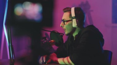 Genç adamın gözlük ve kulaklık takıp mikrofona konuşurken neon ışıkların aydınlattığı stüdyoda podcast yaparken çekilen görüntüsü.
