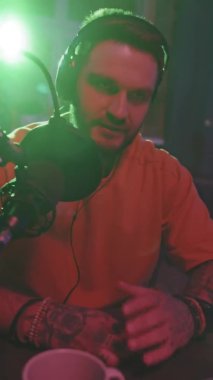 Elinde sakallı, kulaklık takan ve podcast 'te konuşan arka planda yeşil ışığı olan genç bir adamın el kamerası görüntüsü.