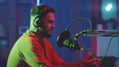 Kulaklık takmış sakallı bir adamın stüdyoda mavi ışıkla oturduğu ve podcast 'te mikrofona konuştuğu yavaş çekim.