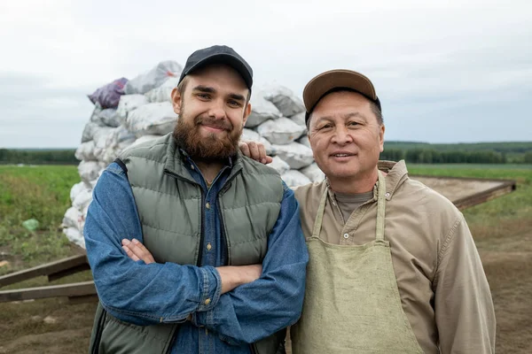 Dwóch szczęśliwych rolników w odzieży roboczej stojących przeciwko stercie worków z ziemniakami — Zdjęcie stockowe