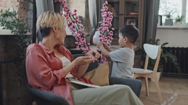 可愛い3歳のアジアの男の子が花びらを裂いて美しい人工の桜の植物を鍋で引き裂き 母親は居心地の良いリビングルームでアームチェアで休んでいる間 — ストック動画