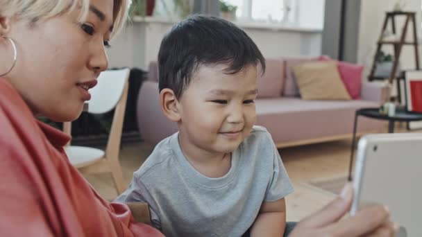 かわいい笑顔の3歳のアジアの男の子が 母親の手にデジタルタブレットのディスプレイを見て面白い顔をして 居心地の良い家で一緒に週末を過ごす — ストック動画
