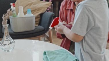 Oturma odasında birlikte ev işi yaparken küçük oğluna temizleyiciyi spreylemeyi öğreten Asyalı bir kadının yavaş çekimleri.