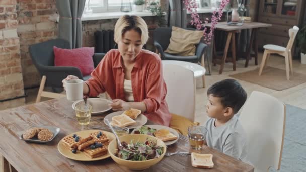 在明亮的现代化公寓里 亚洲母亲和小儿子一起吃早餐的慢镜头在明亮的餐桌边闪烁着 — 图库视频影像
