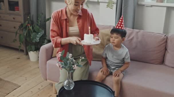 ゆっくりショットの可愛いですアジアの男の子で誕生日の帽子吹きアウトキャンドルオン誕生日ケーキお祝いでお母さんと一緒に居心地の良い家 — ストック動画