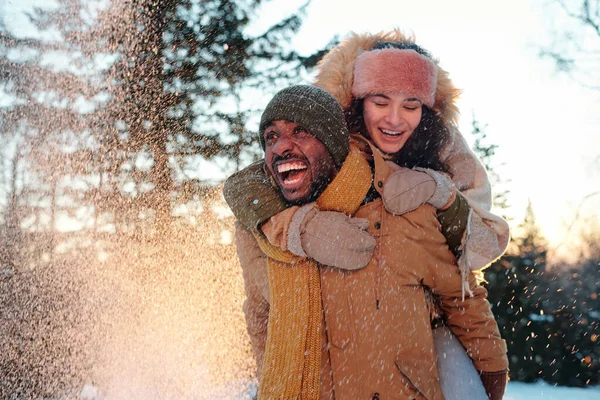 Весела багаторасова пара в зимовому одязі сміється, коли дівчина обіймає свого хлопця — стокове фото