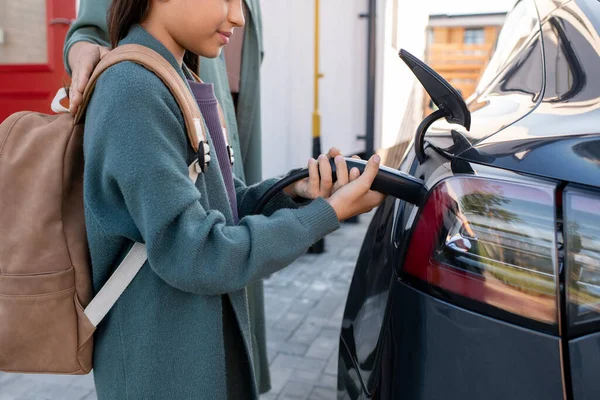 Urocza dziewczyna z plecakiem pomaga ładować samochód elektryczny — Zdjęcie stockowe