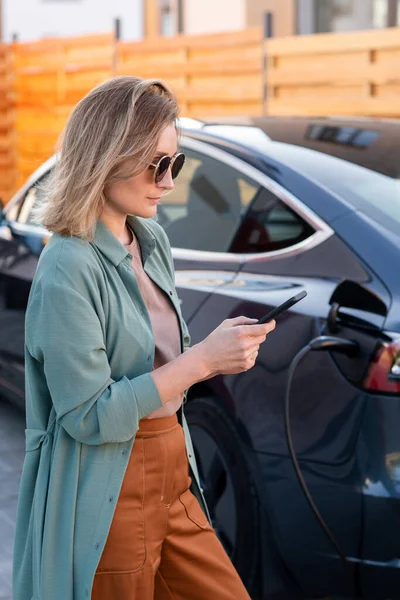 Žena s chytrým telefonem čeká na konec procesu nabíjení vozu — Stock fotografie