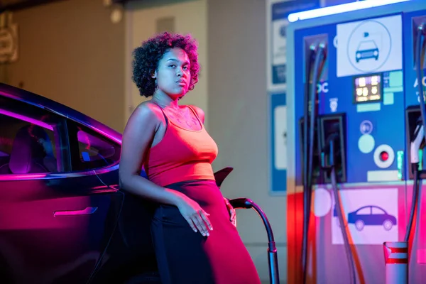 Junge Frau steht mit Elektroauto gegen Tankpfahl — Stockfoto