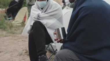 Soğuk bir günde, fakir mülteci kampında battaniyenin altında oturmuş maskeli iki erkek göçmenin yüz maskesi taktığı bir fotoğraf.