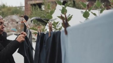 Soğuk bir sabahta göçmen kampına çamaşır asan siyah başörtüsü ve paltolu Müslüman kadının orta ölçekli yakın çekimi.