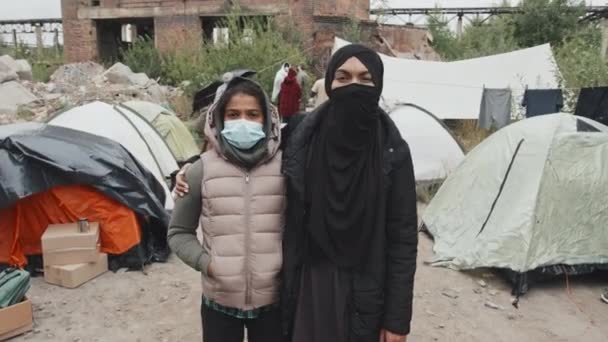穆斯林妇女和她11岁的女儿戴着面具 看着镜头 站在贫穷的难民营里 住在帐篷里的其他移民的中等速度慢画像 — 图库视频影像