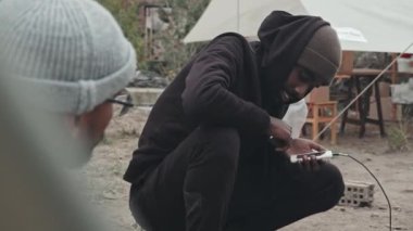 Karma ırk ve Afro-Amerikalı genç mültecilerin orta boy yakın çekimleri. Çadır kentinde, uzatma kablosunun yanında oturan cep telefonlarını kullanıyorlar.