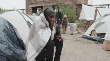 Orta boy, evsiz, Afro-Amerikalı genç bir adam sıcak çay içerken göçmen kampının önünde oturmuş, çocuklu bir anneye merhaba diyor, diğer mültecilerle birlikte yaşıyor.