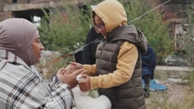 Orta boy bir Afro-Amerikan anne ve küçük kızı dışarıda oturmuş, diğer göçmenlerle birlikte mülteci kampında oynuyorlar.