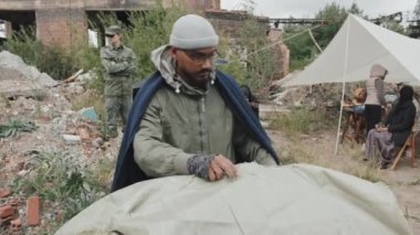 Soğuk bir günde Mülteci kampına çadır kuran sakallı bir adamın diğer göçmenlerle birlikte yaşadığı orta boy bir fotoğrafı. Arka planda duran asker, sakinlerin güvenliğini sağlıyor.
