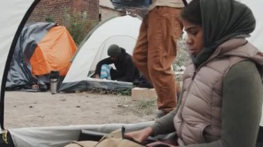 Askeri üniformalı genç erkek polis memuru, diğer göçmenlerle birlikte mülteci kampında çadırda oturan evsiz Afro-Amerikan bir adamla konuşurken not alıyor.