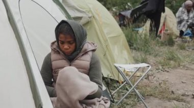 Soğuk rüzgarlı bir günde, elinde battaniyeyle mülteci kampının önünde oturan mutsuz bir Afro-Amerikalı kız resmi.