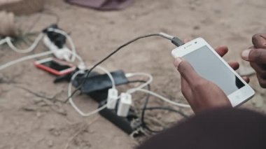 Tanımlanamayan Afro-Amerikan mülteci adamın yakın plan fotoğrafı akıllı telefonuyla kucağında şarj aletleriyle uzatma kablosunun yanında oturuyor.