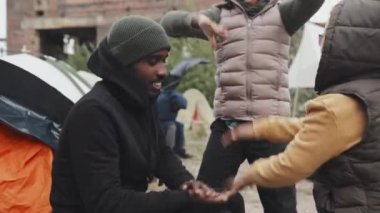 Göçmen kampında çadırlarının önünde iki mülteci kızla oynayan siyah kapüşonlu evsiz genç Afro-Amerikalı adamın orta boy fotoğrafı.