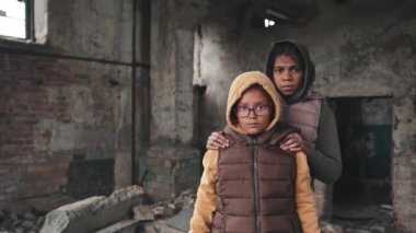 İki Afro-Amerikalı mülteci kızın yıkılmış evlerinin harabelerinde yan yana kameraya bakarken orta boy portresi.