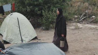Siyah başörtüsü ve paltosu olan Müslüman bir kadının diğer göçmenlerle birlikte yaşadığı mülteci kampının çadırlarında yürürken çekilmiş bir fotoğrafı.