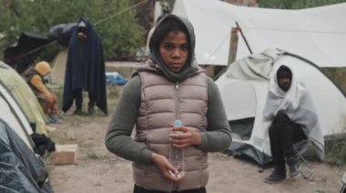 Soğuk bir günde mülteci kampında elinde plastik su şişesiyle duran zavallı Afro-Amerikalı kızın orta boy portresi.