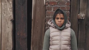 Ortalama yakın plan, üzgün ve yalnız, 11 yaşında, Afro-Amerikalı evsiz bir kız. Açık havada, terk edilmiş ahşap evin karşısında kameraya bakıyor.