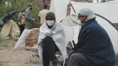 Fakir mülteci kampının dışında battaniyenin altında oturmuş sohbet eden yüz maskeli iki erkek göçmenin orta boy fotoğrafı.