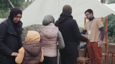 Çadır kentinde yaşayan, yiyecek, su ve giyecek dolu kutular almak için sırada bekleyen çeşitli evsiz ve mültecilerin görüntülerini izliyorum.