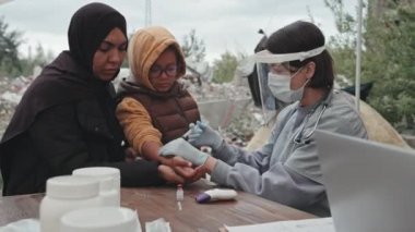 Annesinin yanında duran küçük mülteci kızın belden yukarısında çadır şehrinde yüz koruyucusu takan bir kadın doktor tarafından aşı olurken.