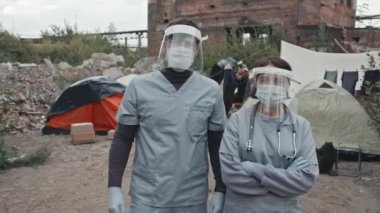 Koruyucu giysiler, yüz kalkanları ve yüz maskeleri takan iki genç tıp çalışanının orta boy portresi mültecilerin ve evsizlerin yaşadığı çadır şehrinde duruyor.