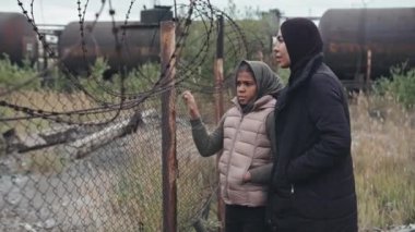 Siyah tesettürlü Müslüman bir kadın ve 11 yaşındaki kızının dikenli tellerin yanında dikilip sohbet ederken orta boy fotoğrafı.